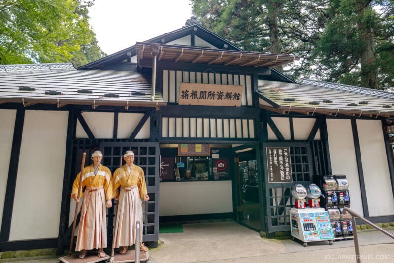 A traditional Japanese building, part of the Hakone Checkpoint Museum, is surrounded by trees, with two mannequins in historical attire at the entrance and vending machines to the right. A sign with Japanese characters hangs above the doorway.