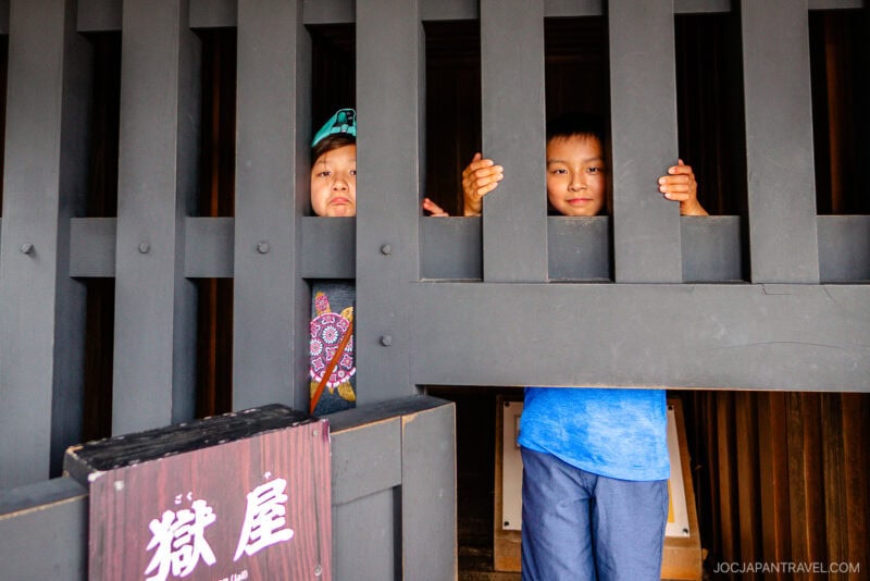Two children stand behind wooden bars at the Hakone Checkpoint Museum, playfully posing as if imprisoned. One child smiles while the other pouts, and a sign with Japanese characters hangs on the bars.