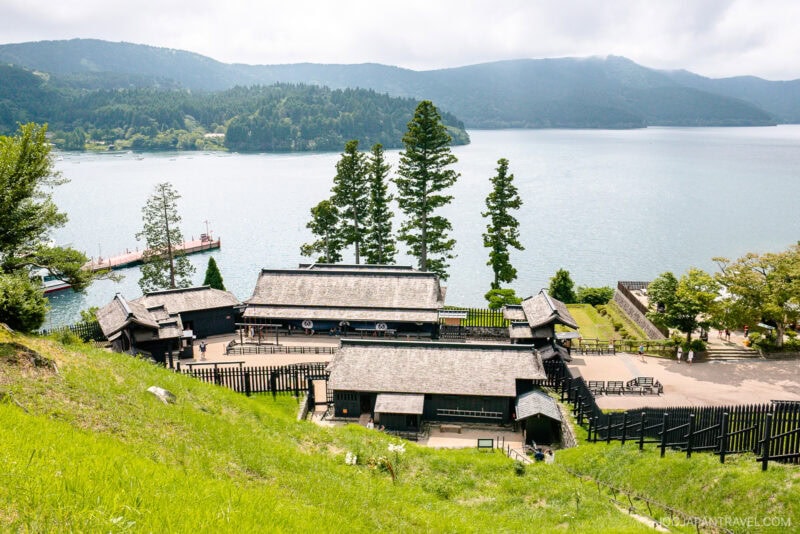 A traditional Japanese building complex with dark wooden roofs sits by a large lakeshore near the Hakone Checkpoint Museum, surrounded by lush greenery and tall trees, with forested hills rising in the distance.