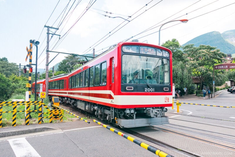 A red and white train, accessible with the Hakone Freepass, crosses a street at a railway crossing surrounded by greenery and mountains, with barriers down and warning lights visible.