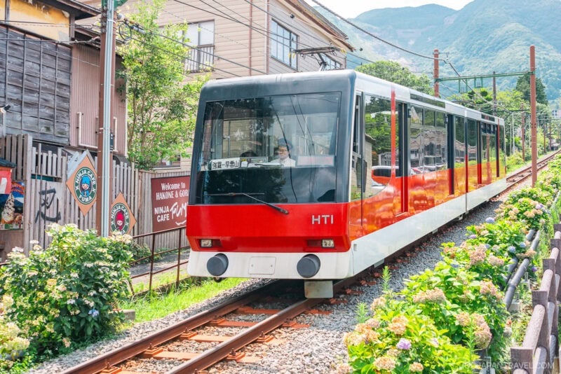 A red and white Hakone Tozan cable car travels up a steep track surrounded by greenery and buildings, with mountains visible in the background&mdash;a scenic ride accessible with the Hakone Freepass.