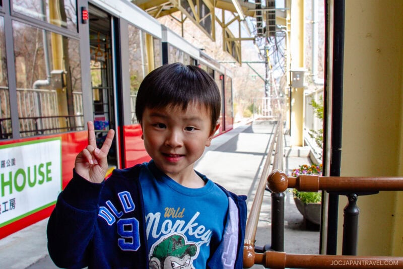 A young boy smiles and holds up a peace sign while standing next to a red and white train at an outdoor station, using his Hakone Freepass. Metal railings line the platform, with trees visible in the background.