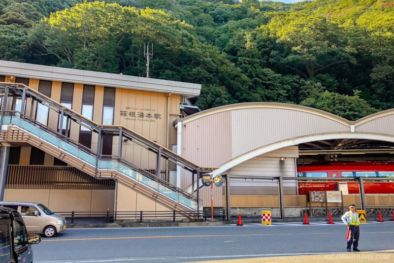 A train station with modern architecture sits at the base of a green hillside. A red train, accessible with the Hakone Freepass, is visible inside, and a uniformed staff member stands near the road in front of the station.