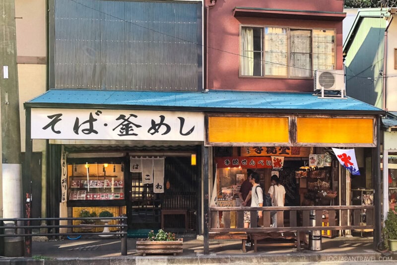 A traditional hakone-yumoto Japanese storefront with a large sign in Japanese characters, wooden details, and two people standing by the entrance; souvenirs and goods are displayed inside.