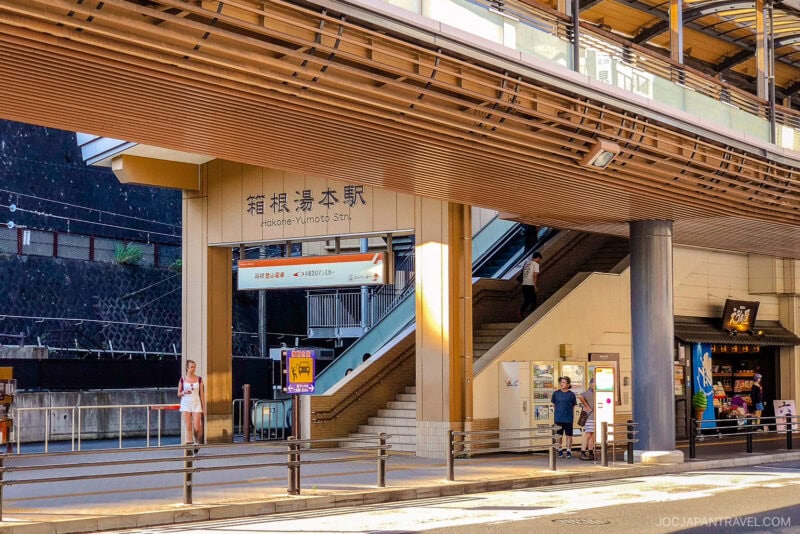 Exterior view of Hakone-Yumoto Station in Japan, with people walking outside, a hakone-yumoto sign in Japanese and English, stairs leading up, and a small shop visible on the right.