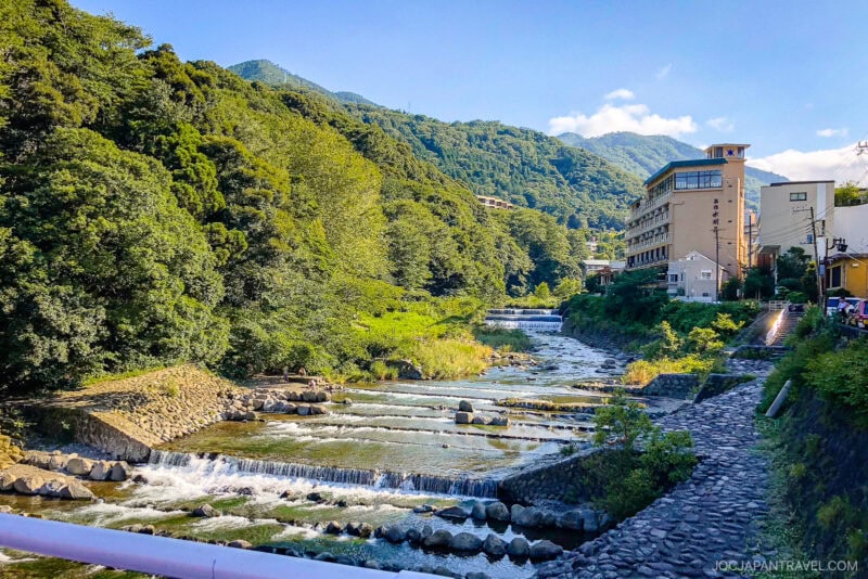 A clear river flows over stone steps between lush green hills and a few buildings in Hakone-Yumoto, with mountains visible in the background under a bright blue sky.