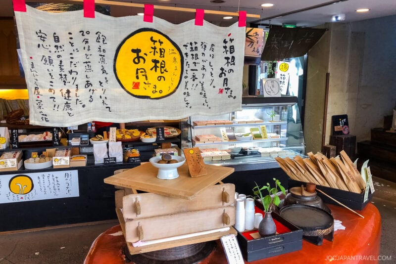 A traditional Japanese sweets shop in Hakone-Yumoto with a large cloth sign featuring Japanese writing, a central table displaying wooden trays, small plants, and a white dessert on a pedestal, plus a refrigerated display case in the background.