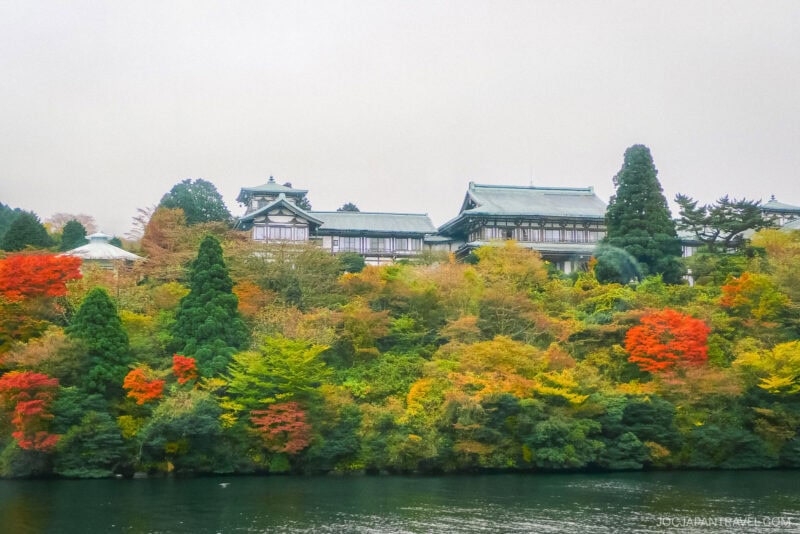 A traditional Japanese building sits atop a hill covered with colorful autumn trees in Hakone-en, overlooking a calm body of water under a cloudy sky.