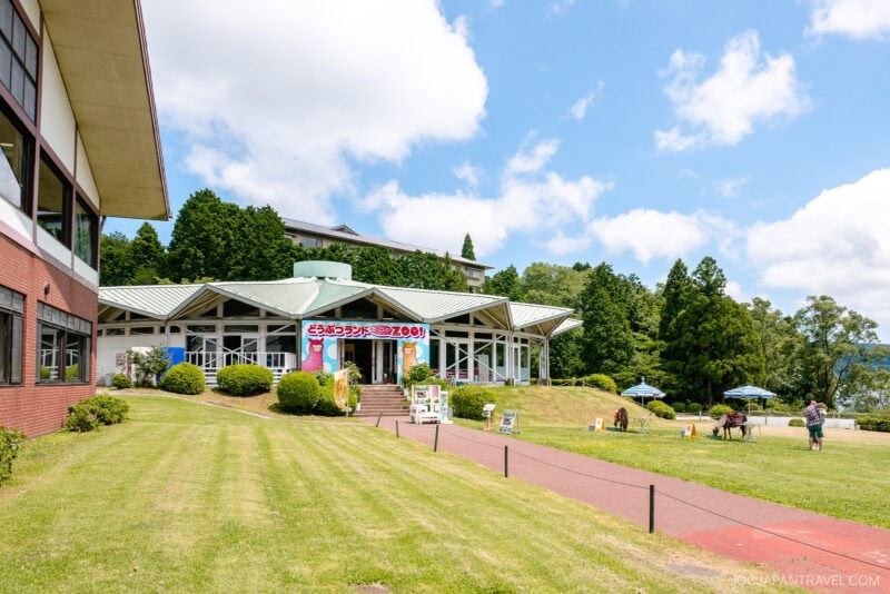 A bright building with colorful cartoon banners sits among green lawns and trees under a blue sky at Hakone-en; people and horses are nearby, and outdoor umbrellas are set up on the grass.