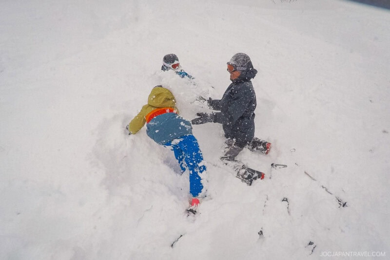 Three people bundled in winter clothing play and laugh together in deep snow, partially buried and surrounded by a snowy landscape during their Hakuba ski trip.
