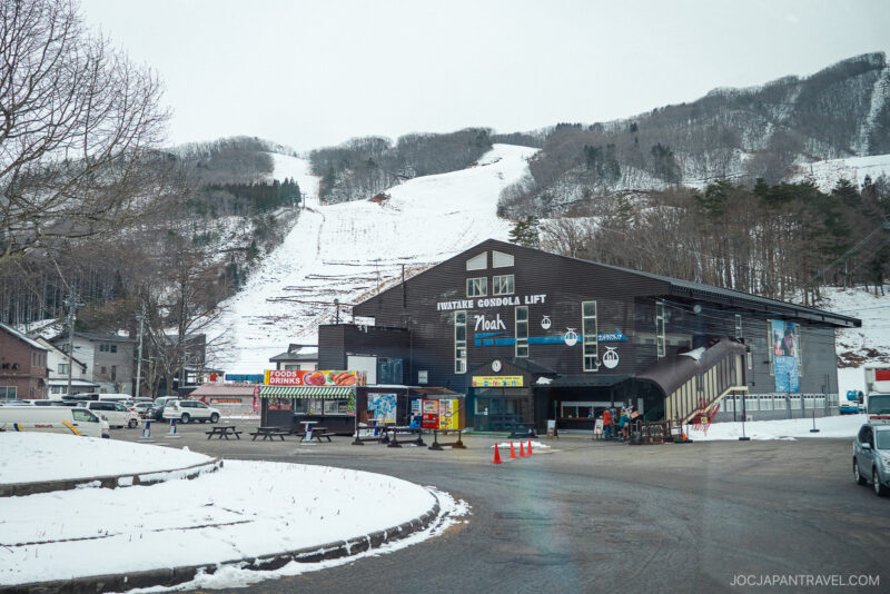 A ski resort entrance during a Hakuba ski trip features a large dark building labeled TWINTAKE GONDOLA LIFT at the base of snowy mountains, surrounded by parked cars, vending stands, and people near the entrance.