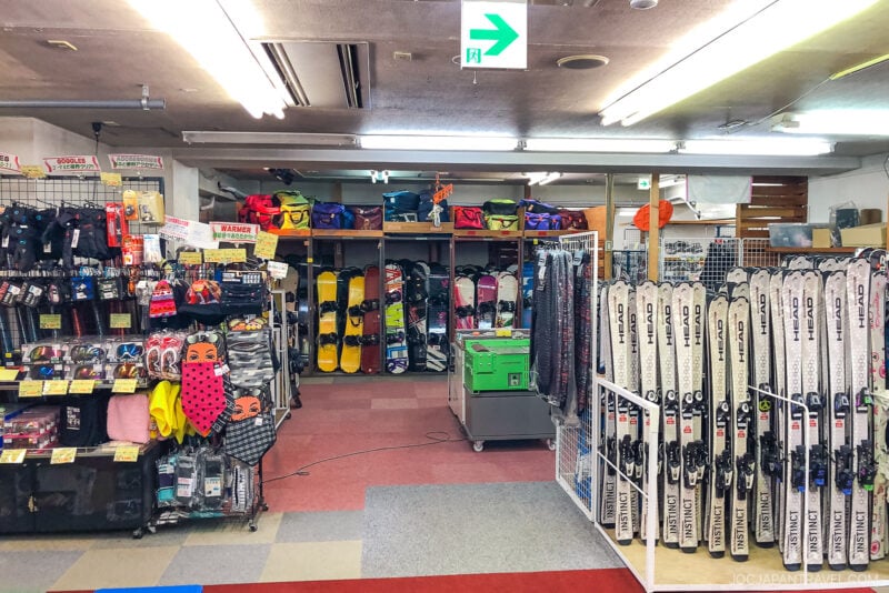 A ski and snowboard rental shop interior perfect for your Hakuba ski trip, with racks of snowboards, skis, helmets, gloves, and winter gear on display. Bright fluorescent lighting and a green emergency exit sign are visible.