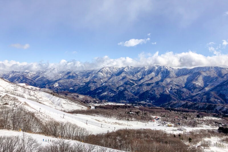 Snow-covered mountains with ski slopes, leafless trees, and small buildings in a valley under a partly cloudy sky. Groups of people are skiing in the foreground, capturing the excitement of a Hakuba ski trip.