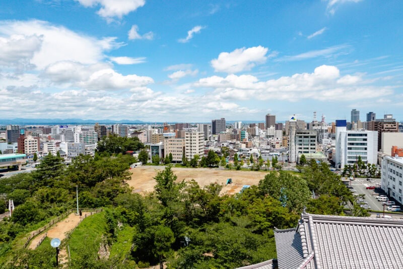 A cityscape view with modern buildings under a blue sky, scattered clouds, and a mix of green trees and open land in the foreground, with Hamamatsu Castle rising gracefully amidst the urban landscape.