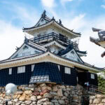 Hamamatsu Castle (浜松城) – Home of Tokugawa Ieyasu and the Castle of Success A traditional Japanese castle, like Hamamatsu Castle, with white walls and black tiled roofs, stands atop a stone foundation beneath a blue sky with light clouds. Some trees and a few people are visible near the entrance.
