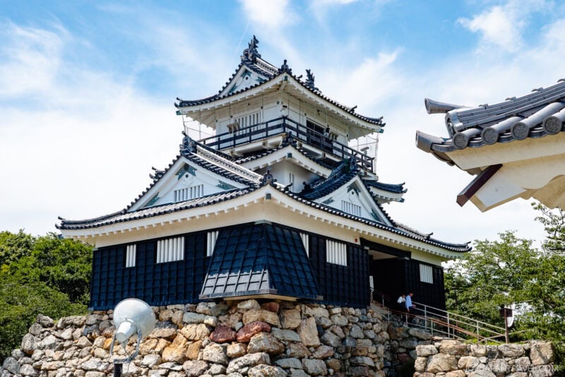 A traditional Japanese castle, like Hamamatsu Castle, with white walls and black tiled roofs, stands atop a stone foundation beneath a blue sky with light clouds. Some trees and a few people are visible near the entrance.