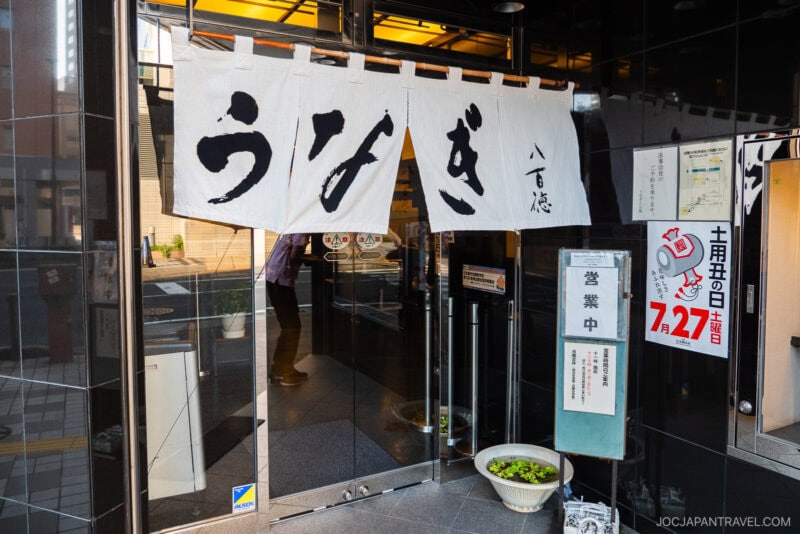 A Japanese restaurant entrance with white noren curtains displaying large black kanji, glass doors, potted plants, and various signs—including one highlighting a hamamatsu unagi special event on July 27.