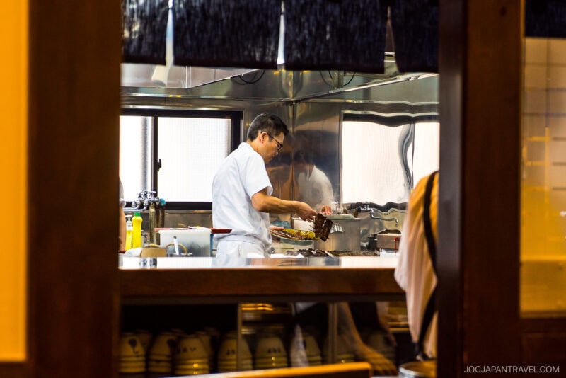 A chef in a white uniform prepares food in a restaurant kitchen, seen through a wooden window frame. Cooking utensils, plates, and fresh hamamatsu unagi are visible on the counter.