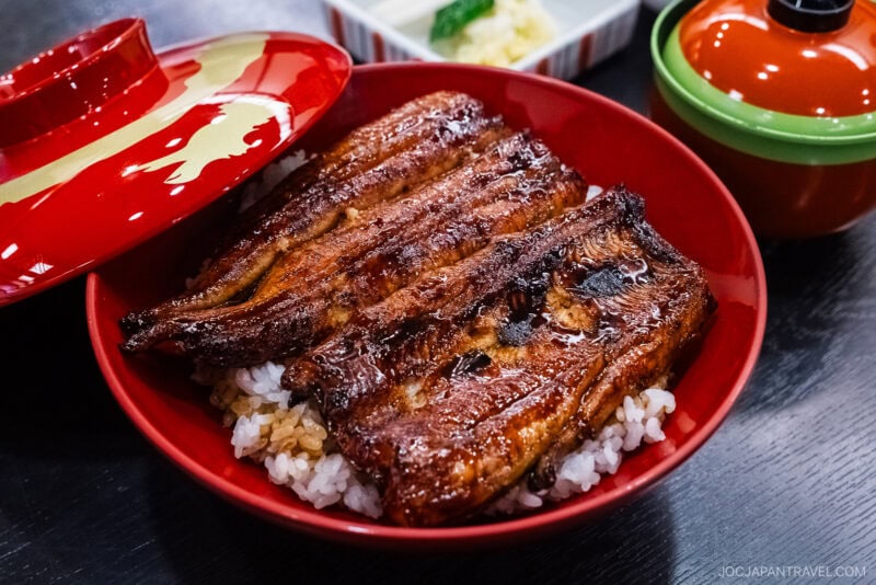 A red bowl filled with hamamatsu unagi—grilled eel glazed with sauce—served over steamed white rice. There is also a red and gold lid, a green-lidded bowl, and a small side dish with pickles in the background.