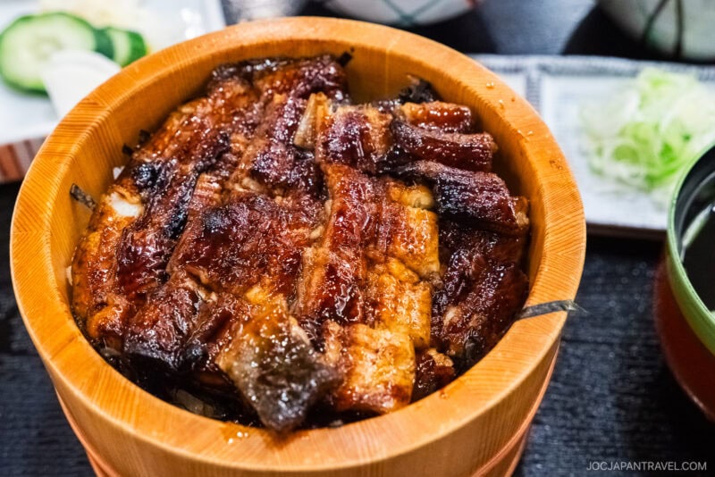 A close-up of a wooden bowl filled with Hamamatsu unagi&mdash;grilled eel glazed in sauce over rice&mdash;accompanied by cucumber slices, sliced green onions, and soup in the background.