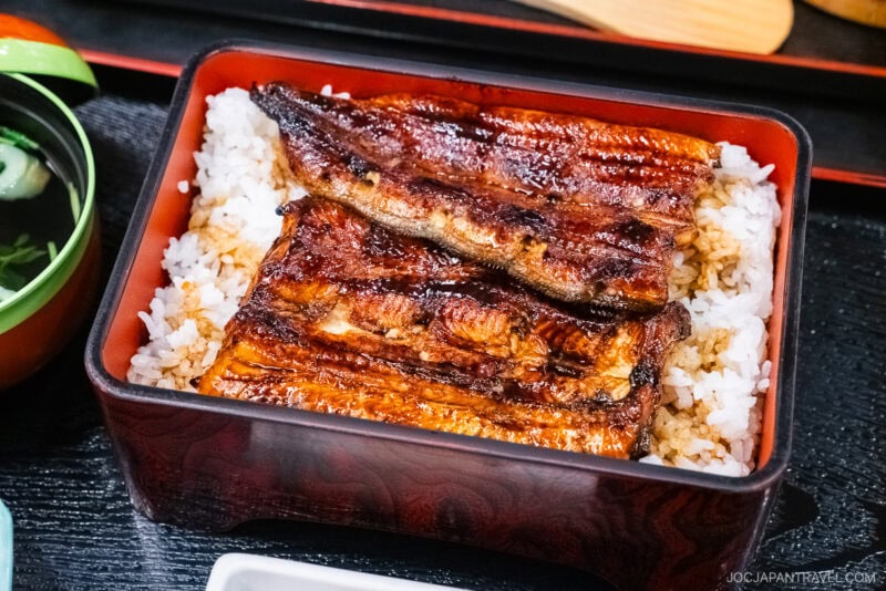 A lacquered box filled with white rice topped with grilled, glazed hamamatsu unagi fillets, served alongside a small bowl of soup.
