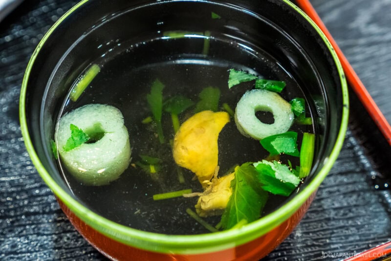 A close-up of a bowl of clear Japanese soup with green herbs, two round pieces of fish cake, and a yellow mushroom, served in a lacquered bowl on a dark tray—a perfect starter before savoring Hamamatsu unagi.