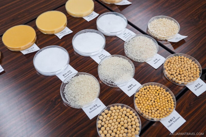 Clear petri dishes display samples of rice, soybeans, and powders on a dark wood table. Each dish is labeled, showcasing ingredients used in miso, much like those seen on a hikari miso factory tour and arranged in neat rows.