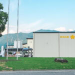Hikari Miso Factory Tour A white industrial building with HIKARI MISO written in yellow stands on green grass, with flagpoles in front and mountains visible in the background—a perfect spot to begin your hikari miso factory tour under a blue sky.