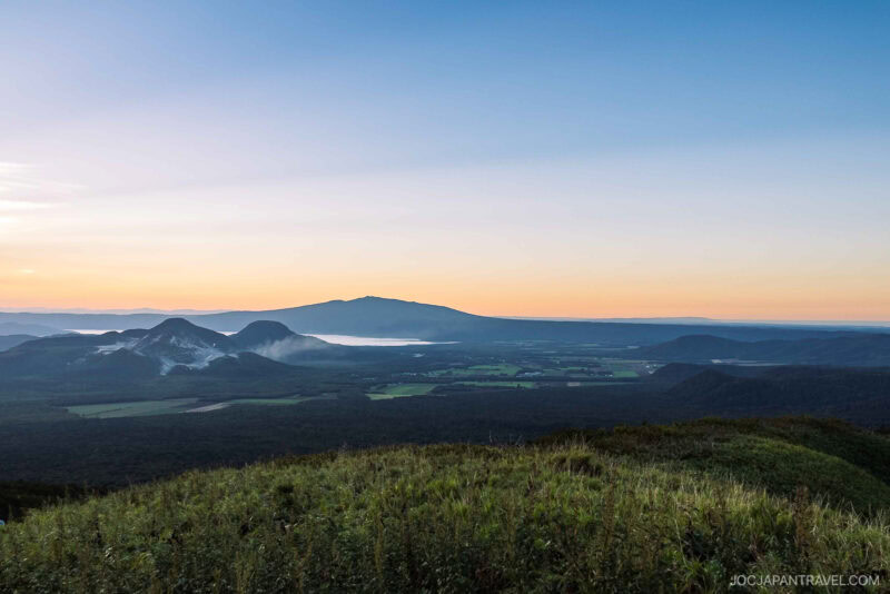 A scenic sunrise view of a vast green landscape with distant mountains, misty valleys, and the serene beauty of Lake Akan and Lake Mashu under a clear sky gradually brightening on the horizon.