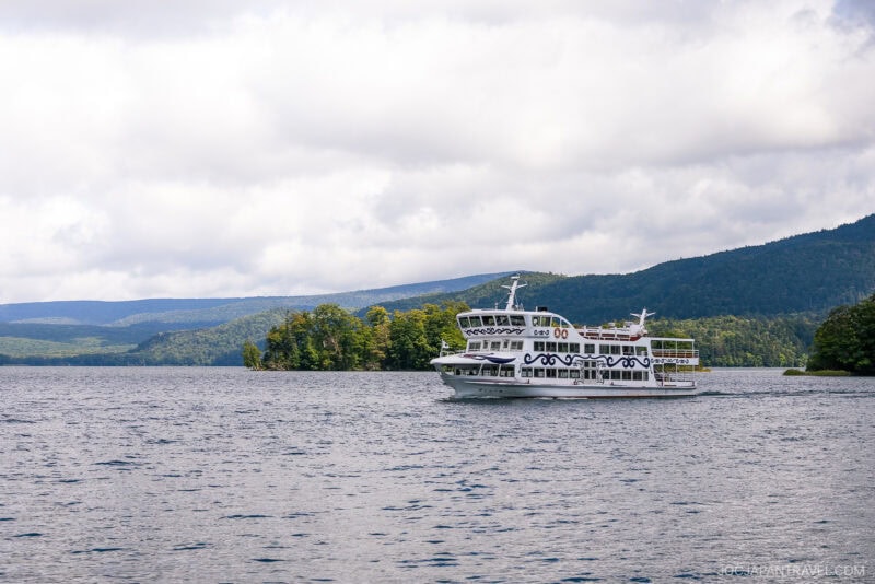 A white sightseeing boat cruises on calm Lake Akan, surrounded by green, forested hills under a cloudy sky.
