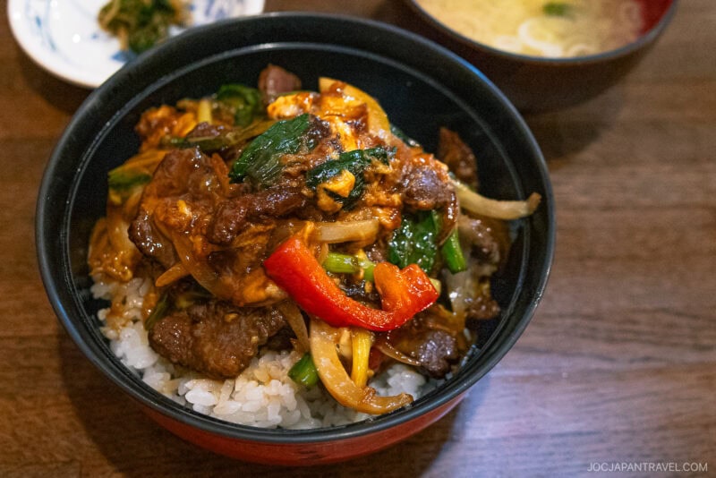 A bowl of rice topped with stir-fried beef, red bell pepper, onions, and leafy greens evokes the fresh flavors found near Lake Akan and Lake Mashu. A small bowl of soup and a side dish are partially visible in the background on a wooden table.
