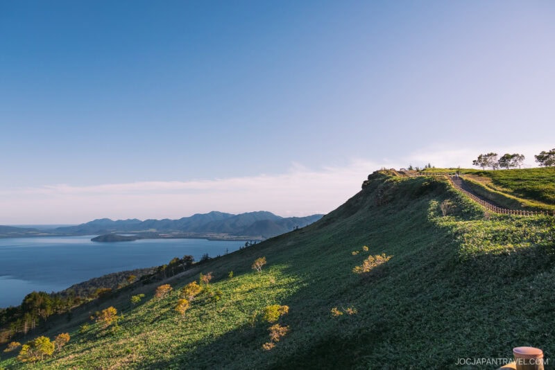 A sunlit green hillside overlooks the calm waters of Lake Akan, with distant mountains under a clear blue sky, while a path winds along the hill’s edge, lined with small trees.