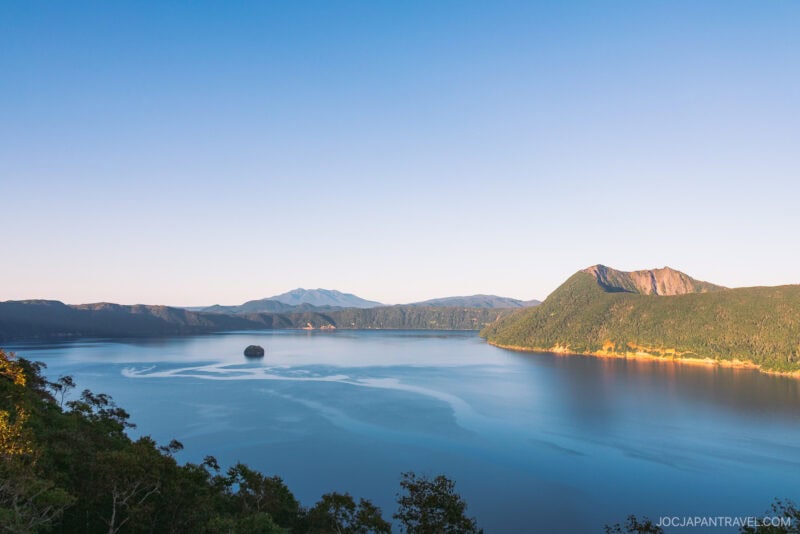 A serene lake, reminiscent of Lake Akan or Lake Mashu, is surrounded by forested hills and mountains under a clear blue sky. Sunlight casts a warm glow on the landscape, with a small island visible near the center of the calm water.