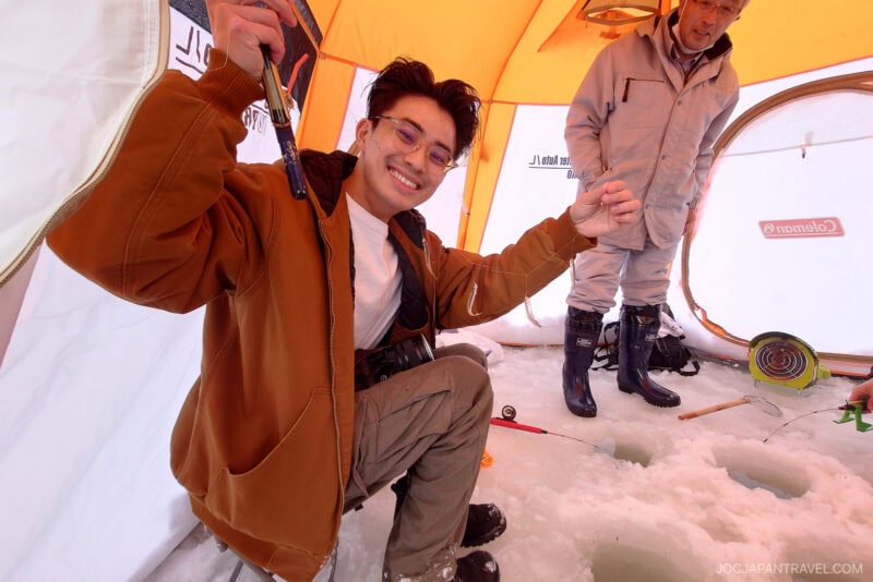A smiling person in a brown jacket holds up a small fish inside an ice fishing tent in Mikasa City, while another stands nearby on the snowy ground with fishing gear and ice holes.