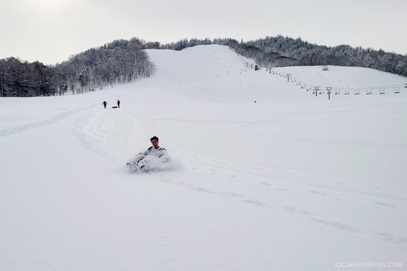 A person sleds down a snow-covered slope in Mikasa City, leaving clear tracks behind. The snowy landscape and distant hills frame the scene, while a few people walk in the background beneath a cloudy sky.