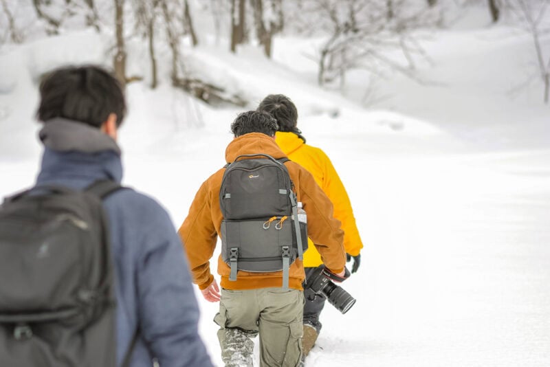 Three people wearing backpacks and winter jackets walk in a line through deep snow in a snowy, wooded area in Mikasa City. One person carries a camera in their right hand, with leafless trees visible in the background.