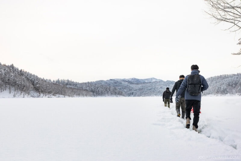Four people walk in a single file line through deep snow across a frozen, snow-covered landscape near Mikasa City, surrounded by trees and distant mountains under a cloudy sky.