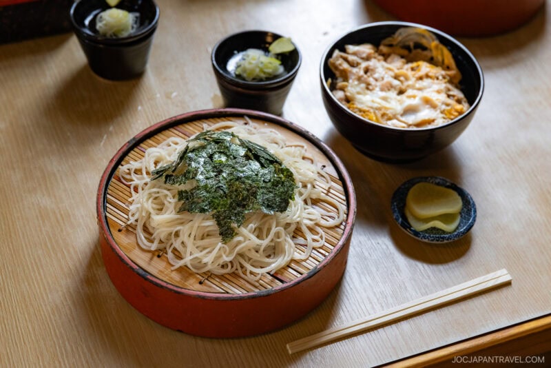 Enjoy a Japanese meal in Mikasa City, featuring a bamboo tray of cold soba noodles topped with seaweed, a bowl of rice with meat and egg, small bowls of pickles, and dipping sauce, with chopsticks on the table.