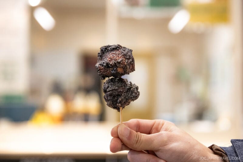 A hand holds a wooden skewer with two pieces of dark, crispy fried food, possibly meat, against a blurred indoor background in Mikasa City.