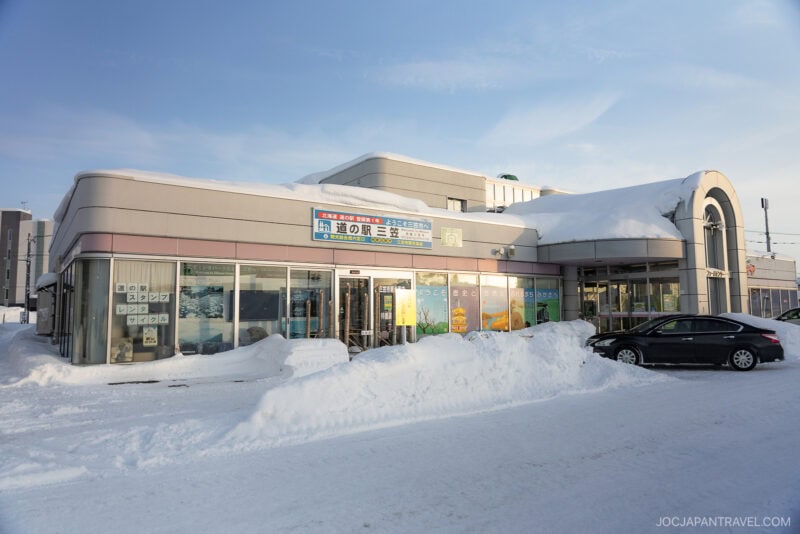 A modern building in Mikasa City is covered in snow, with large windows and Japanese signs above the entrance. Snow piles up around the walkway, and a black car is parked in front under a clear blue sky.