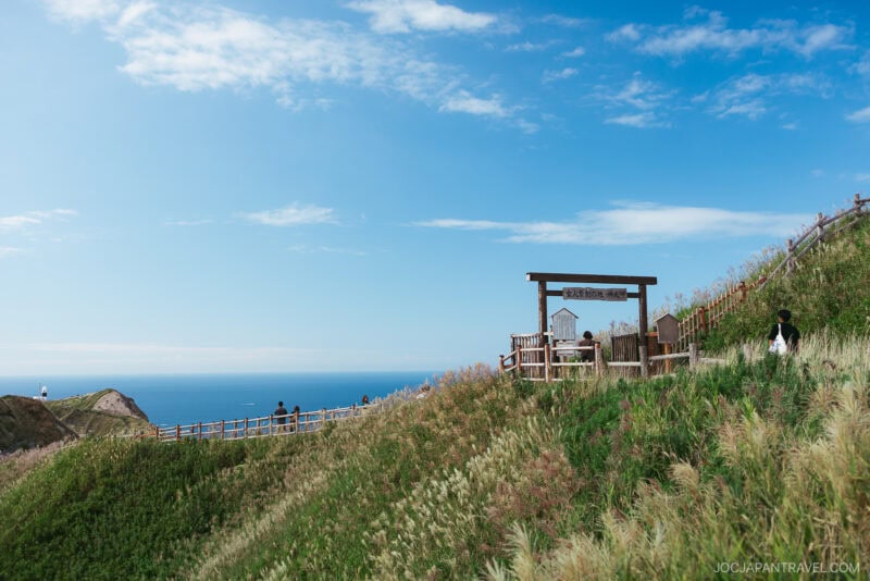 A wooden viewing platform and fence overlook the grassy Yoichi hills, with a bright blue sky and ocean in the background. A few people walk along the path, enjoying the scenic landscape.