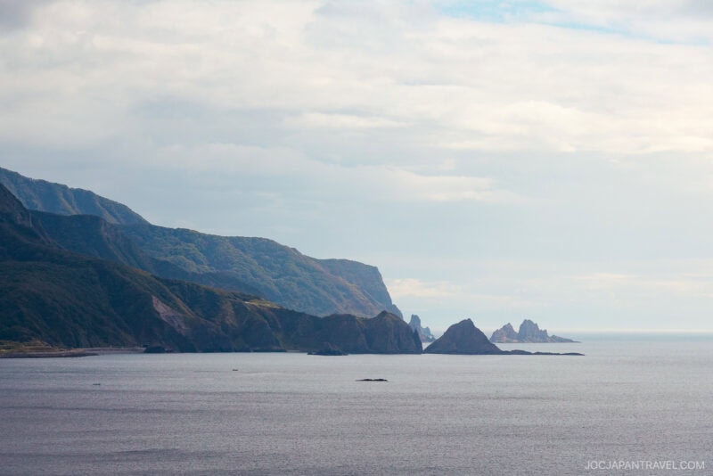 A tranquil coastal landscape near Yoichi, with steep green hills, rocky outcrops, and calm blue water under a partly cloudy sky. The coastline stretches into the distance, creating a peaceful and scenic view.