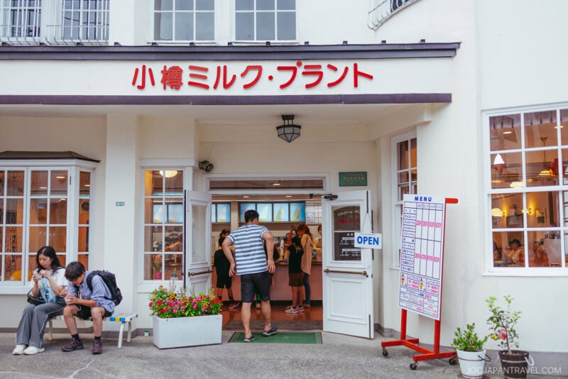 People stand outside and inside a white building with Japanese signage above the entrance in Yoichi. Some sit on a bench eating, while others enter or exit the shop. A menu board and OPEN sign are visible near the doorway.
