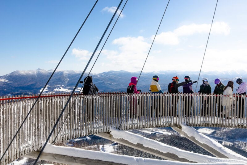 A group of people in winter clothing stand on a snow-covered lookout platform with safety cables at Hoshino Resorts Tomamu, overlooking a mountain landscape under a clear blue sky.