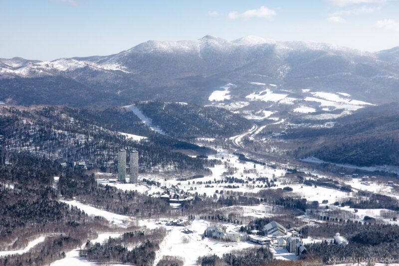A snowy mountain landscape with tall, forested hills and distant peaks. In the valley below, Hoshino Resorts Tomamu’s buildings and tall towers are scattered among snow-covered trees under a partly cloudy sky.