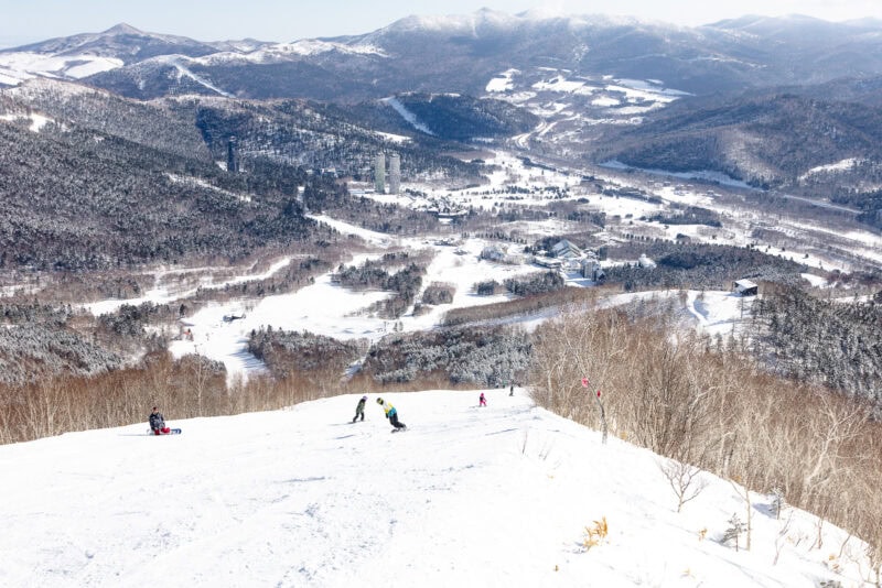 Skiers and snowboarders descend a snowy slope at Hoshino Resorts Tomamu, with a wide view of the mountain landscape, snow-covered trees, and distant buildings under a clear sky.