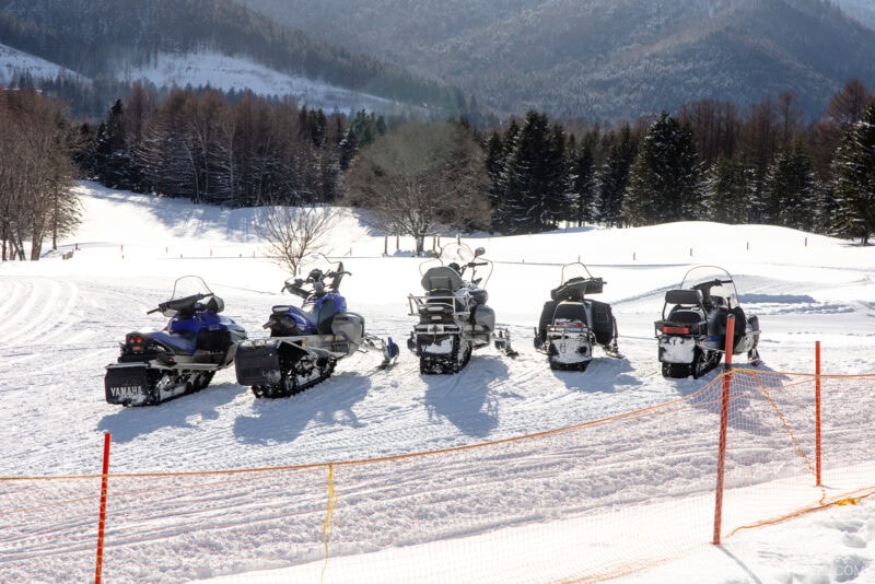 Five snowmobiles are parked side by side on a snowy field at Hoshino Resorts Tomamu, with trees and mountains in the background on a sunny winter day. An orange safety net runs along the foreground.