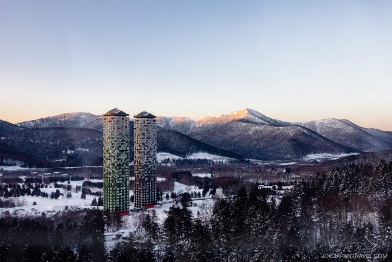 Two tall, colorful towers of Hoshino Resorts Tomamu rise amid a snowy landscape dotted with pine trees, framed by mountain ranges under a clear sky at sunrise or sunset.