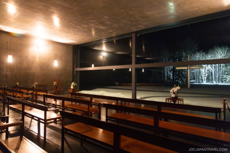 A modern chapel at Hoshino Resorts Tomamu features wooden benches, soft wall lights, and large windows overlooking a snowy landscape with trees illuminated at night.