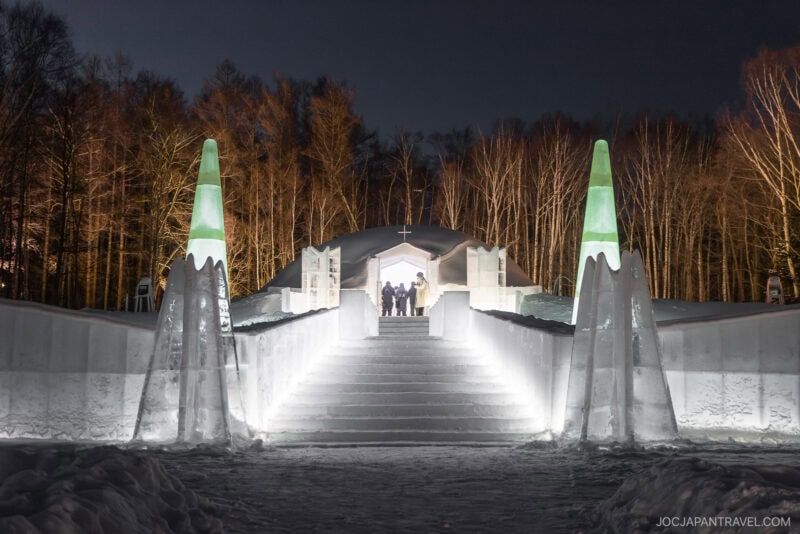 An illuminated ice and snow structure at Hoshino Resorts Tomamu features stairs leading to an arched entrance, flanked by two tall, green-tipped ice towers, set against leafless trees under the night sky.